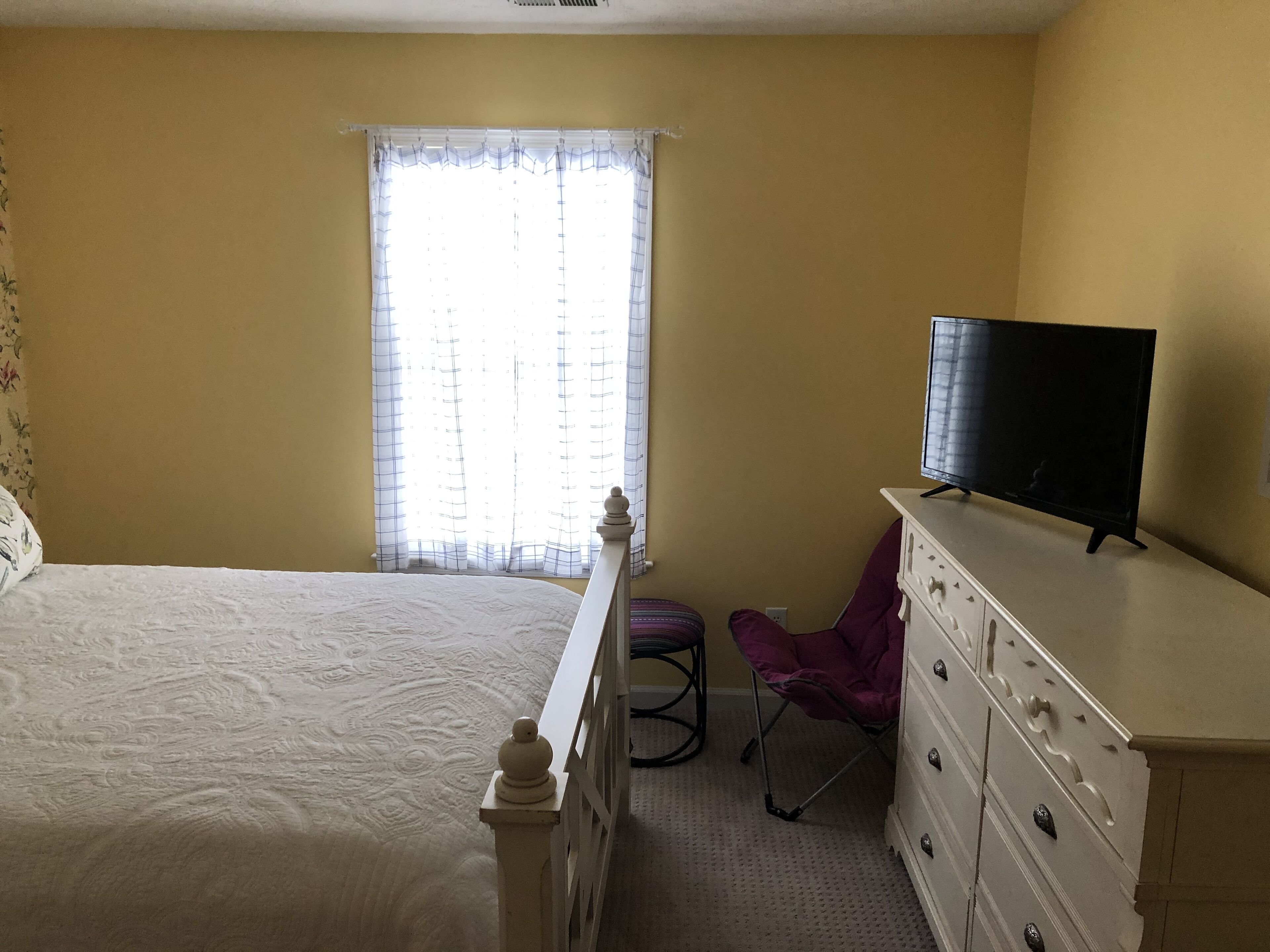 Un dormitorio con cama, cómoda, televisor y ventana con cortinas traslúcidas.
