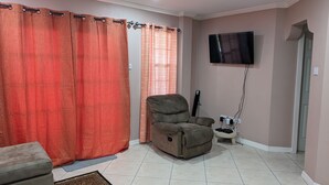 A living room with a sofa, a recliner, a flat-screen TV mounted on the wall, and a small table with a power strip. - Malay Getaway and taxi service (Laborie)