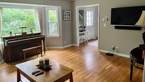 Un salon avec un piano, un téléviseur fixé au mur et une vue sur l’extérieur grâce à de grandes fenêtres.