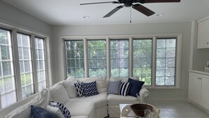 Un salon moderne avec un canapé d’angle blanc, une table basse et un ventilateur de plafond. De grandes fenêtres laissent entrer la lumière naturelle et offrent une vue sur la verdure environnante.