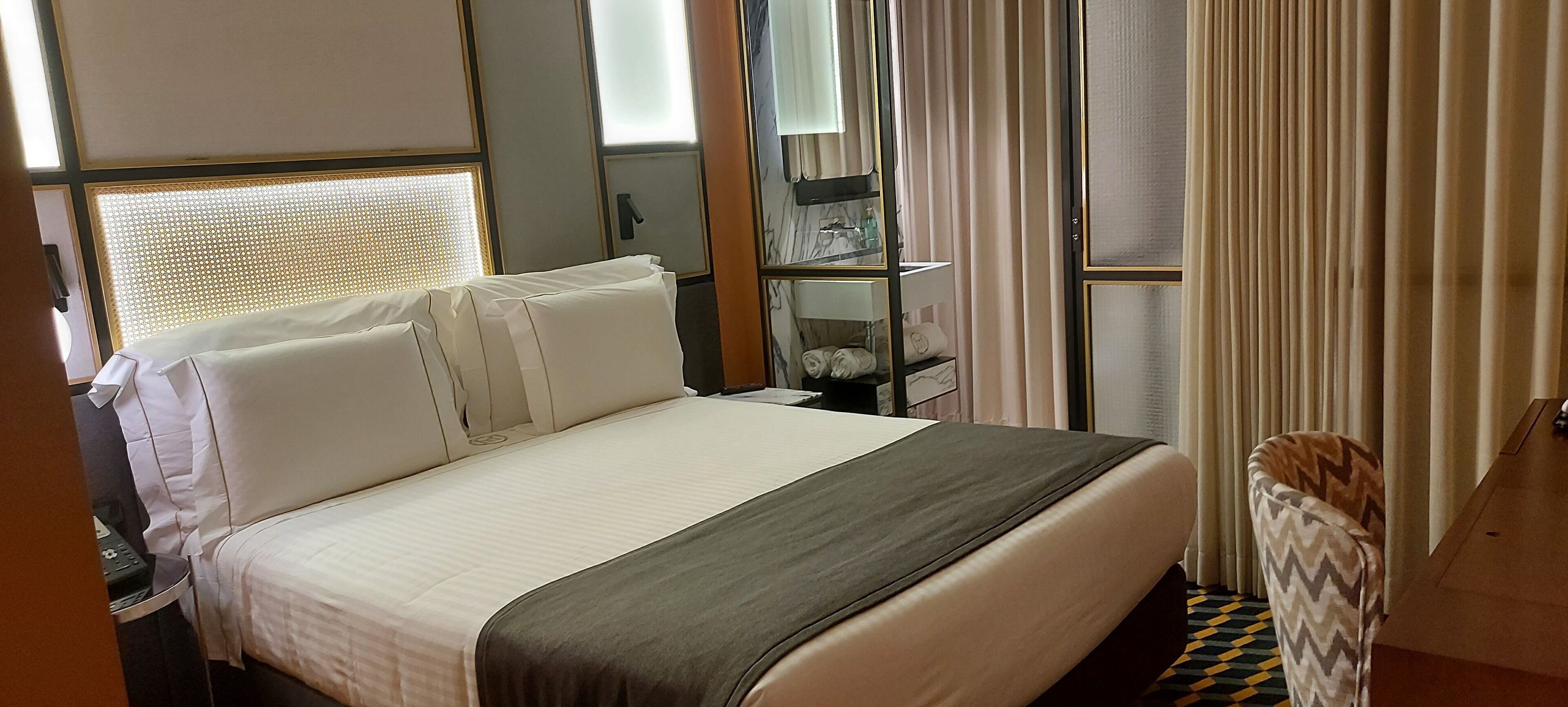 A hotel room with a large bed, a bedside table, a mirror, and a window with curtains.
