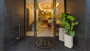 Ingresso moderno di un hotel con porta in vetro, tappetino nero con logo e due piante in vaso.