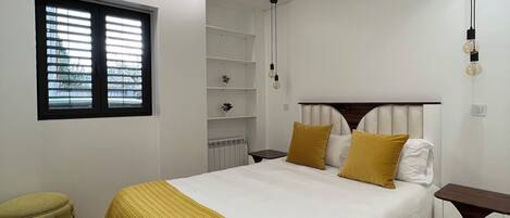 A modern bedroom with a large bed, a bedside table, a small stool, and a window with blinds.