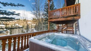 A hot tub on a wooden deck with a view of snow-covered trees and buildings. - Meadows Townhome (Avon)