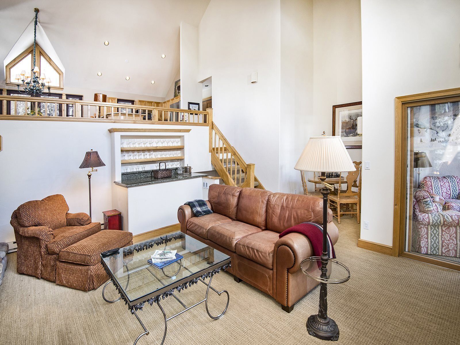 A spacious living room with a brown leather sofa, a glass coffee table, and a patterned armchair. A staircase with wooden railings leads to an upper level.