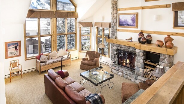 A cozy living room with a stone fireplace, large windows, and a high ceiling. - Meadows Townhome (Avon)