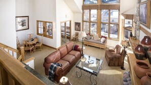 A spacious living room with a high ceiling, large windows offering a view of the outdoors, a brown leather sofa, a glass coffee table, and a stone fireplace. - Meadows Townhome (Avon)
