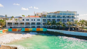 A multi-story hotel with balconies, a beach in front, and palm trees. - Maho Beach House - Spacious 1 Bedroom Suite (Maho, Sint Maarten)