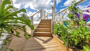 A wooden deck leading to a staircase with a white railing, surrounded by greenery and a clear sky. - Maho Beach House - Spacious 1 Bedroom Suite (Maho, Sint Maarten)