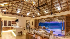 A spacious living area with a large window overlooking a beach, a wooden ceiling, and a kitchen area with modern appliances. - Oceanfront Luxury Property in Elite Development (Huatulco)