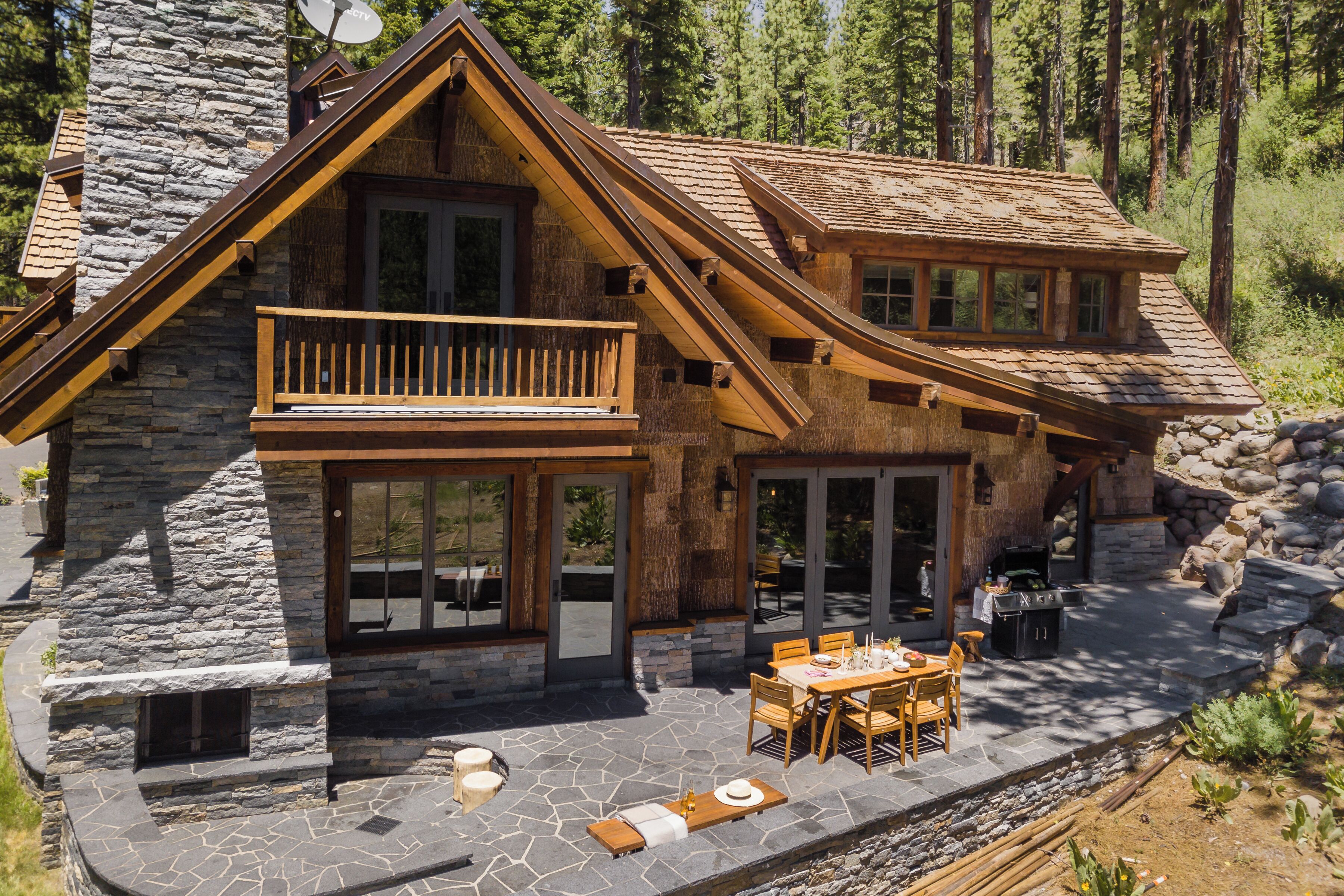 Une cabane en rondins à deux étages, avec une façade en pierre, un balcon et un espace patio aménagé avec du mobilier d’extérieur.