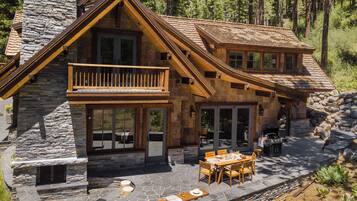 Une cabane en rondins à deux étages, avec une façade en pierre, un balcon et un espace patio aménagé avec du mobilier d’extérieur.