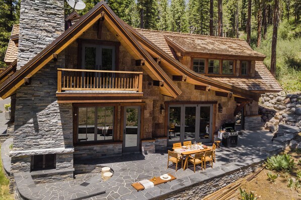 Una cabaña de madera de dos pisos con fachada de piedra, un balcón y un patio con mobiliario exterior.