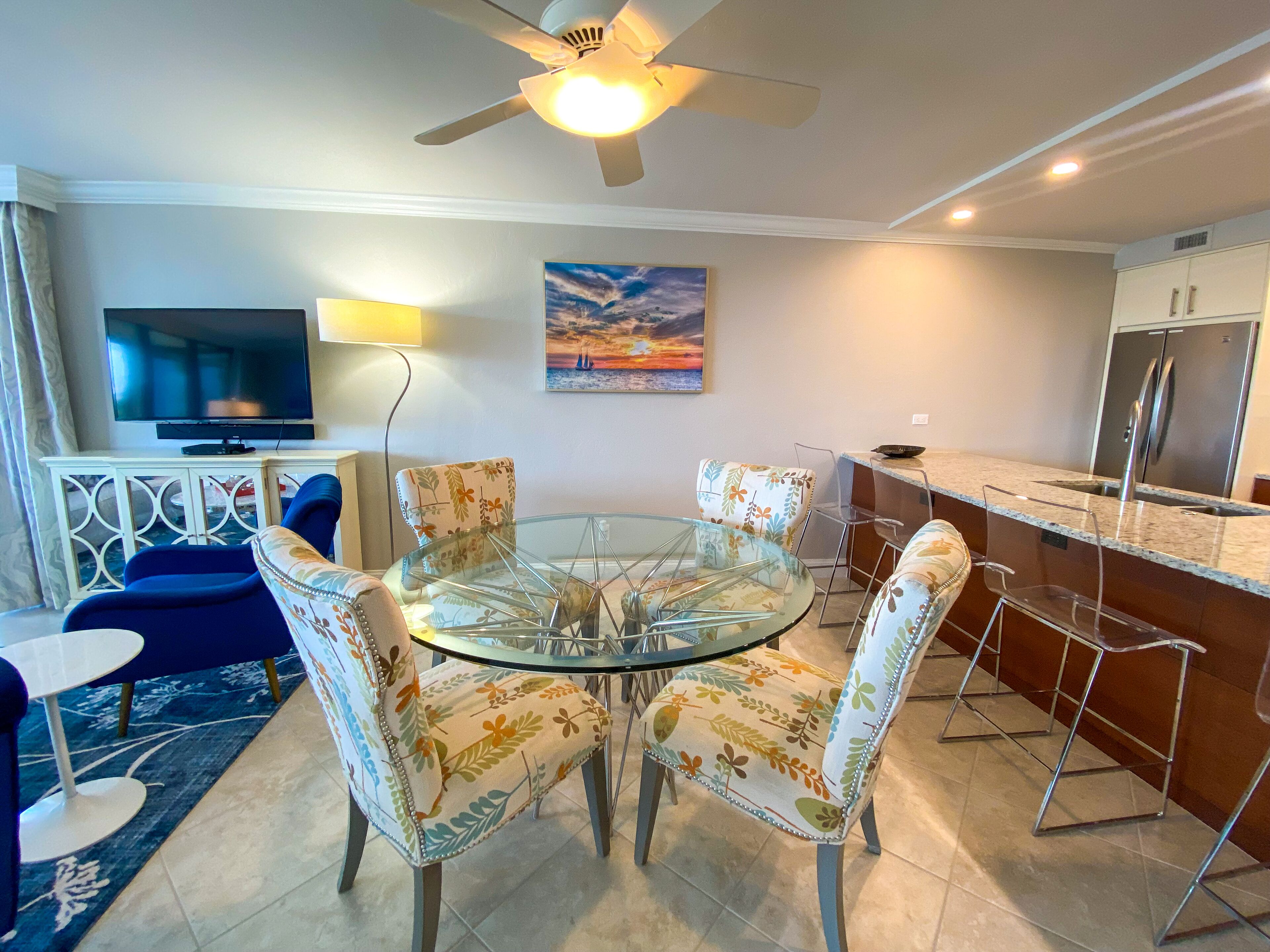A dining area with a glass table surrounded by floral-patterned chairs, a TV on a cabinet, a painting on the wall, and a kitchen with a refrigerator.