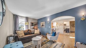 Ein modernes Wohnzimmer mit einer Couch, Sesseln und einem Couchtisch. Durch einen Bogen ist eine Küche erkennbar.