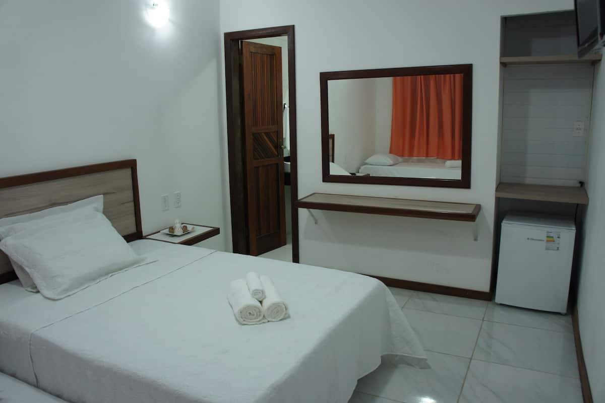 A hotel room with a bed, a nightstand, a mirror, a shelf, and a small refrigerator.