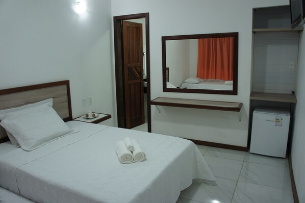 A hotel room with a bed, a nightstand, a mirror, a shelf, and a small refrigerator. - Pousada Camburi (Cairu)