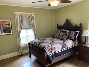 A bedroom with a wooden bed, a nightstand with a lamp, a window with curtains, and a ceiling fan.