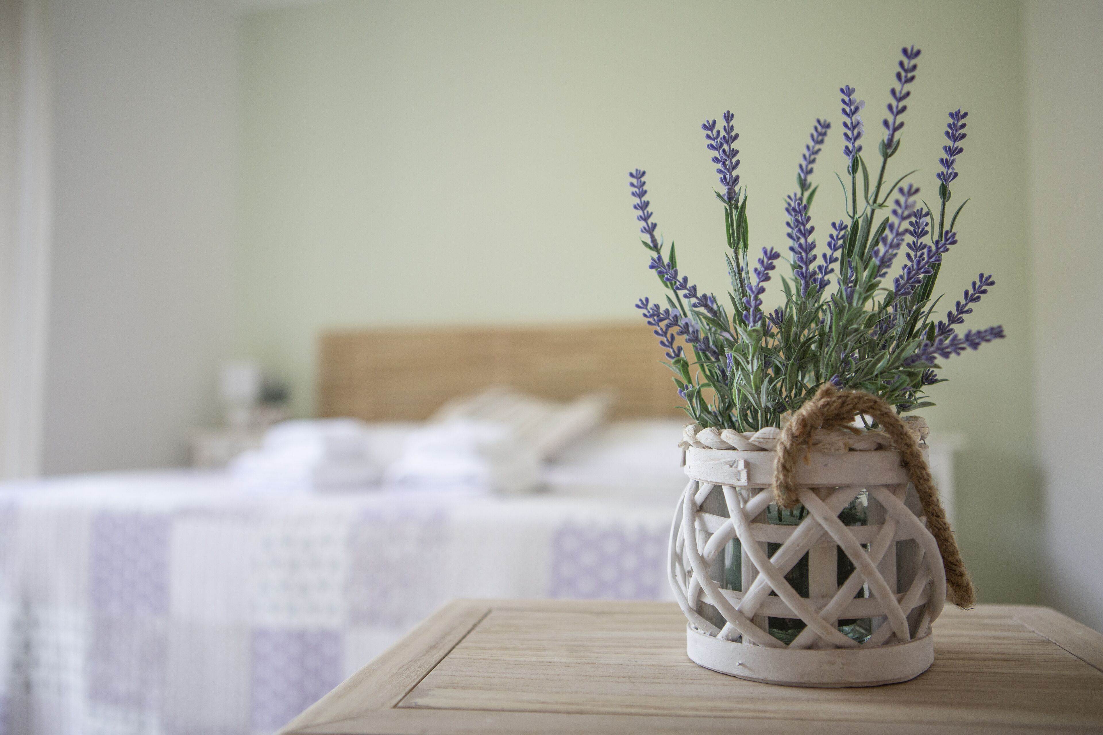 Une lanterne blanche en osier, ornée de plantes à la lavande, est posée sur une table en bois dans une chambre à coucher qui contient un lit et une tête de lit blanche.