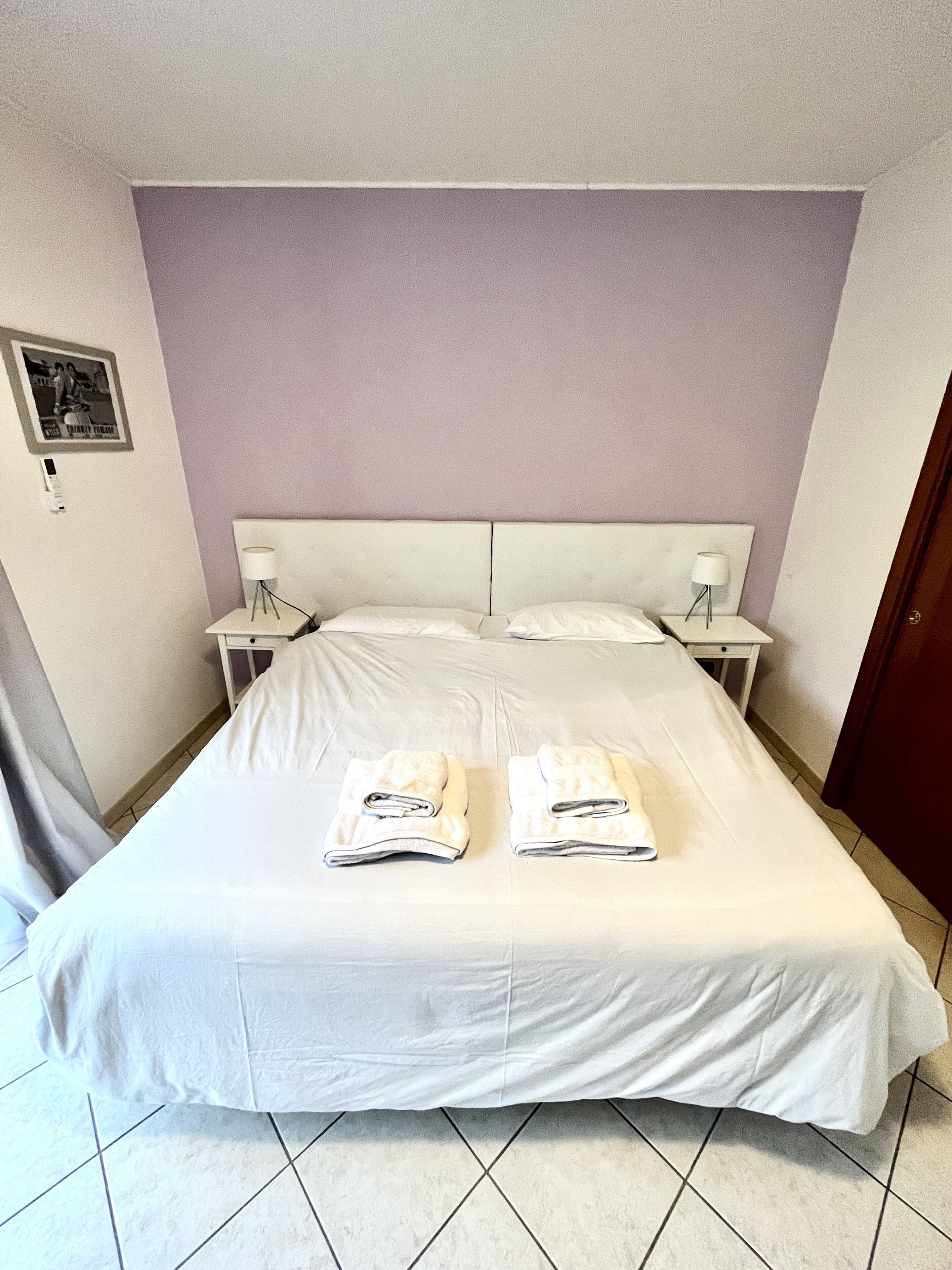 A neatly made bed with two folded towels, a white headboard, and two bedside tables with lamps.