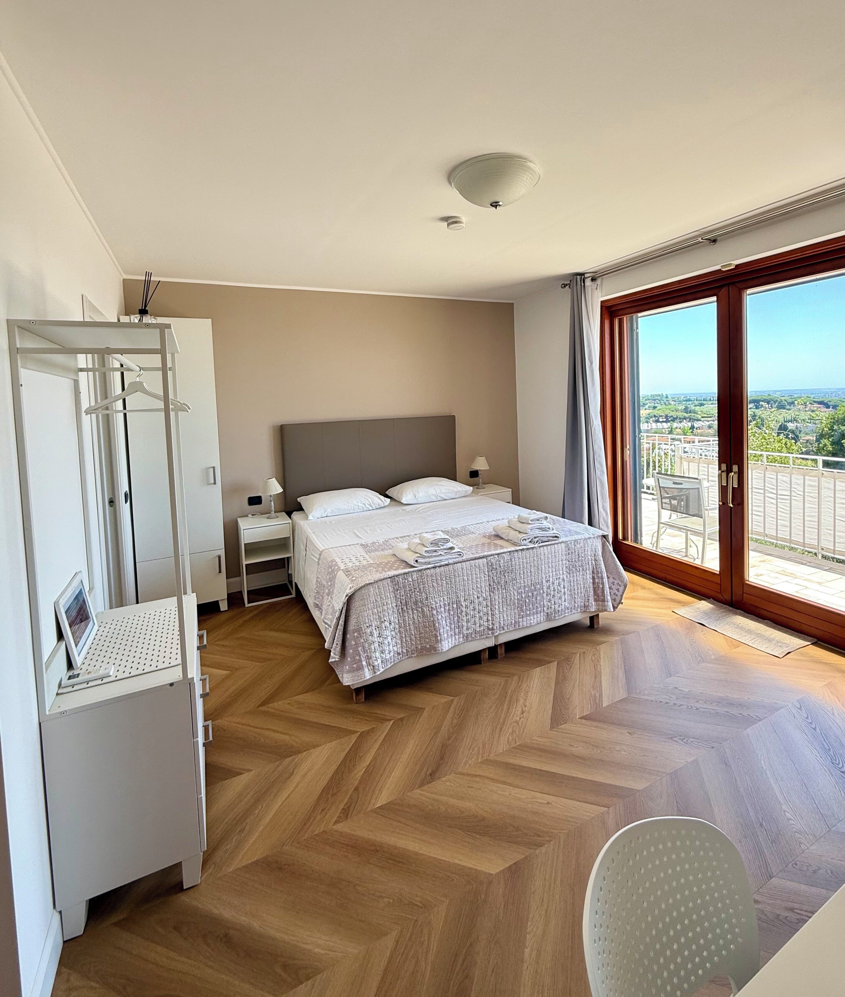 A bedroom with a bed, bedside table, lamp, wardrobe, and a view through a large window.