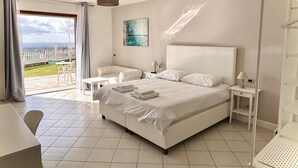 A modern bedroom with a large bed, a white nightstand, a chair, and a table. There is a view of the outdoors through a glass door. - Cape House (Albano Laziale)