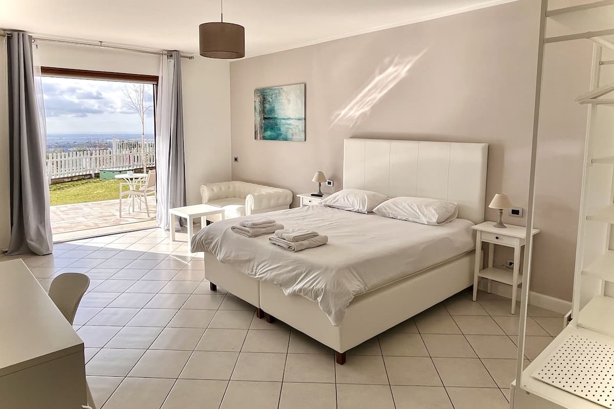 A modern bedroom with a large bed, a white nightstand, a chair, and a table. There is a view of the outdoors through a glass door.