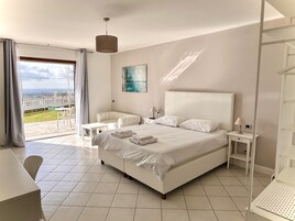 Un dormitorio moderno con una cama grande, una mesita de noche blanca, una silla y una mesa. Se ve el exterior a través de una puerta de vidrio.