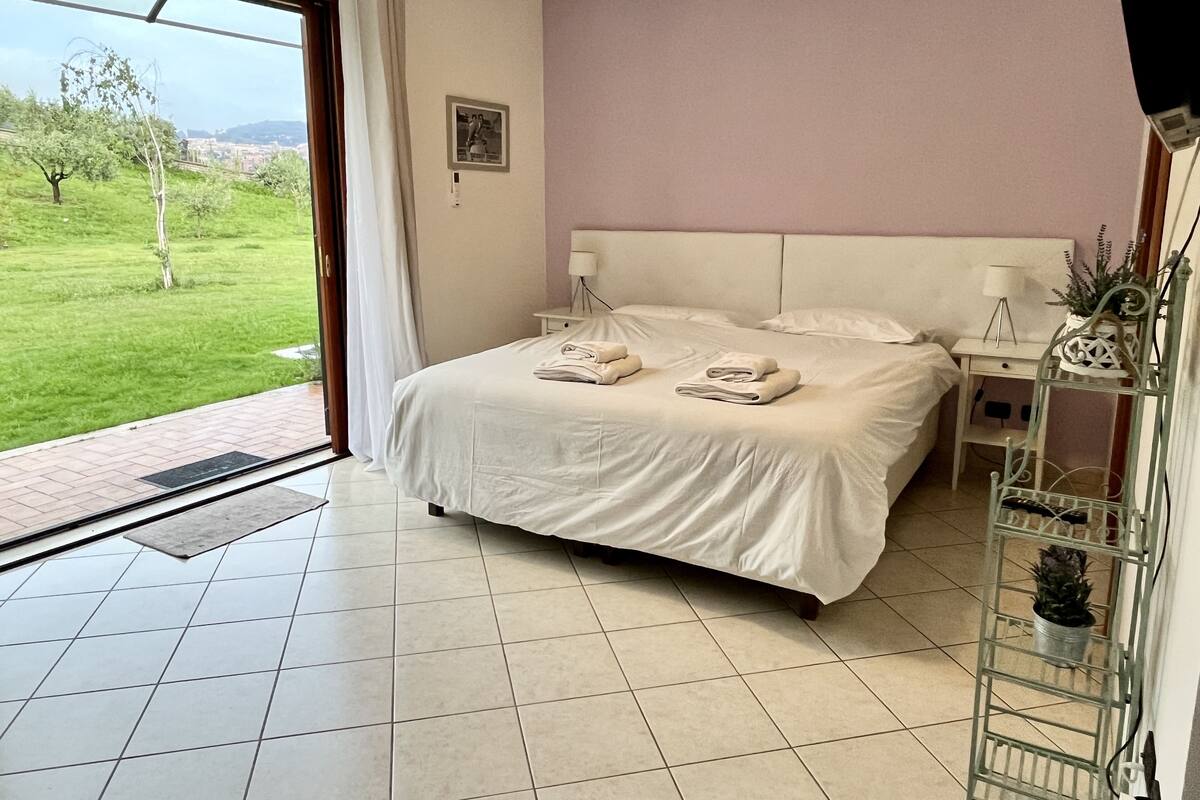 A bedroom with a bed, a nightstand, a lamp, and a view of a green landscape.