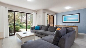A living room with a grey sofa, a coffee table, and a TV on a blue brick wall. - Bay Parklands, 57/2 Gowrie Ave - ground floor unit with pool, tennis court & aircon (Nelson Bay)