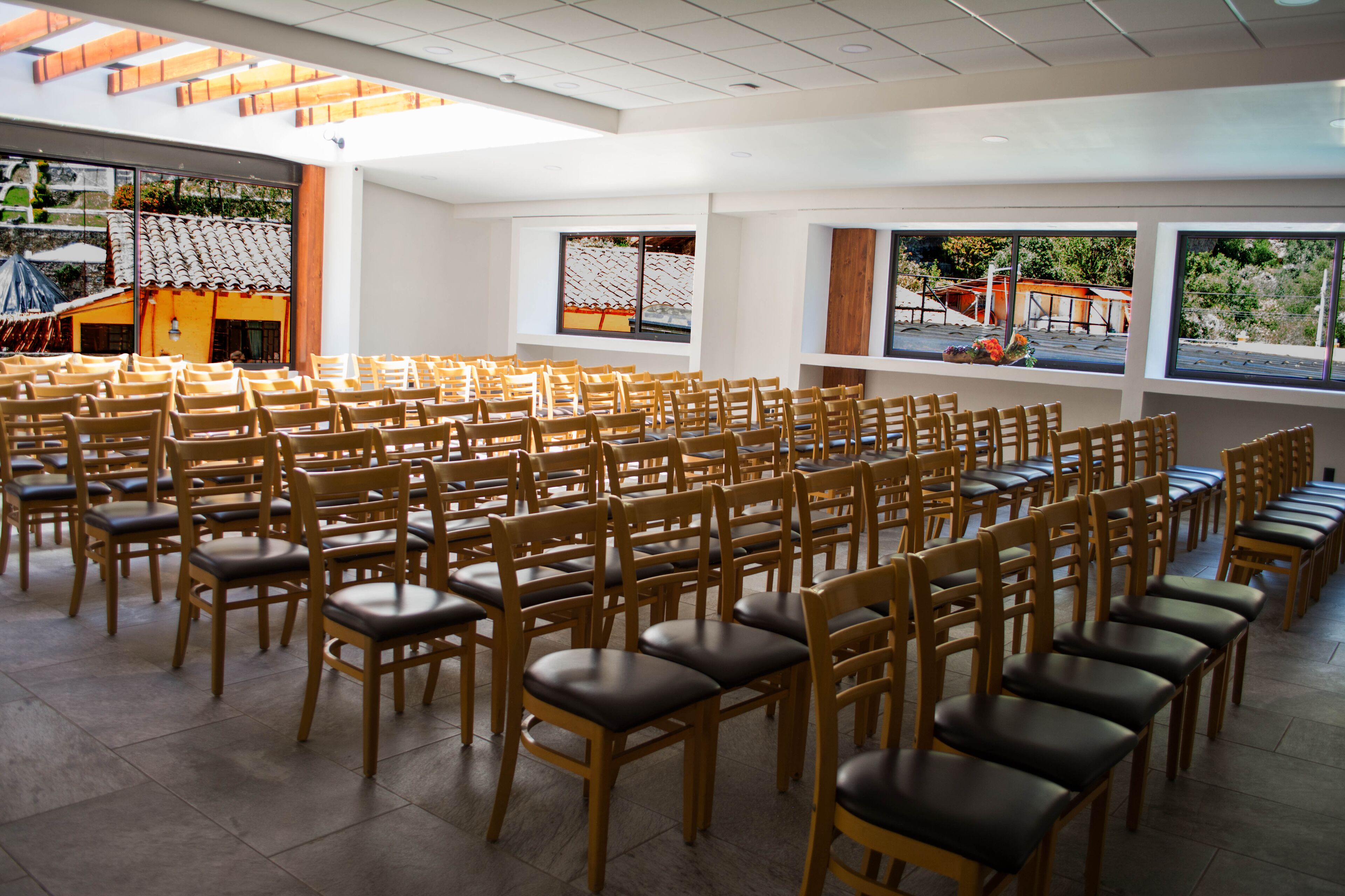 A spacious hall with rows of wooden chairs and tables, large windows with views of the outdoors, and a modern interior design.