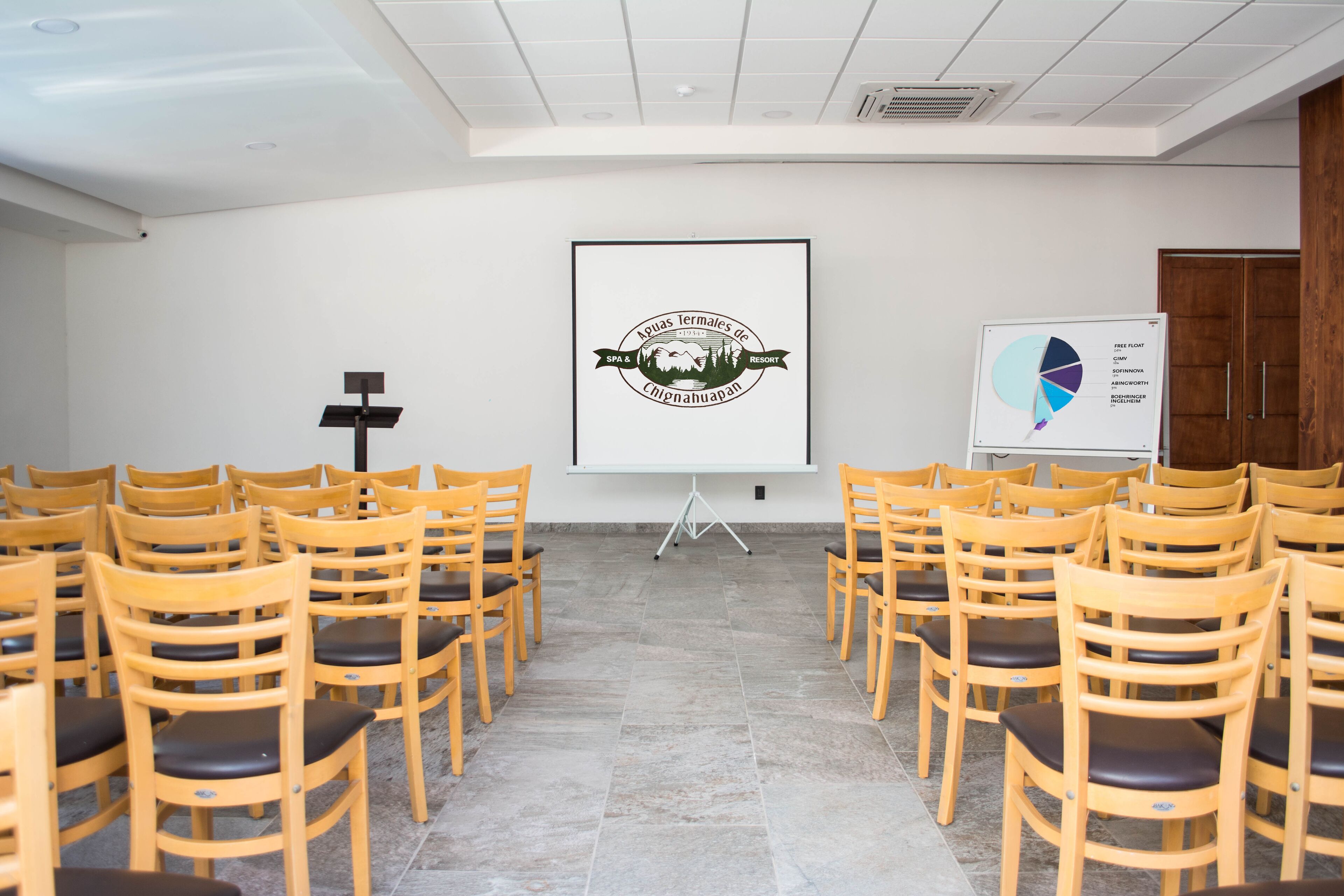 Ein Konferenzraum mit Reihen aus Holzsesseln, einer Projektionsleinwand und einer Tafel mit einem Kreisdiagramm.