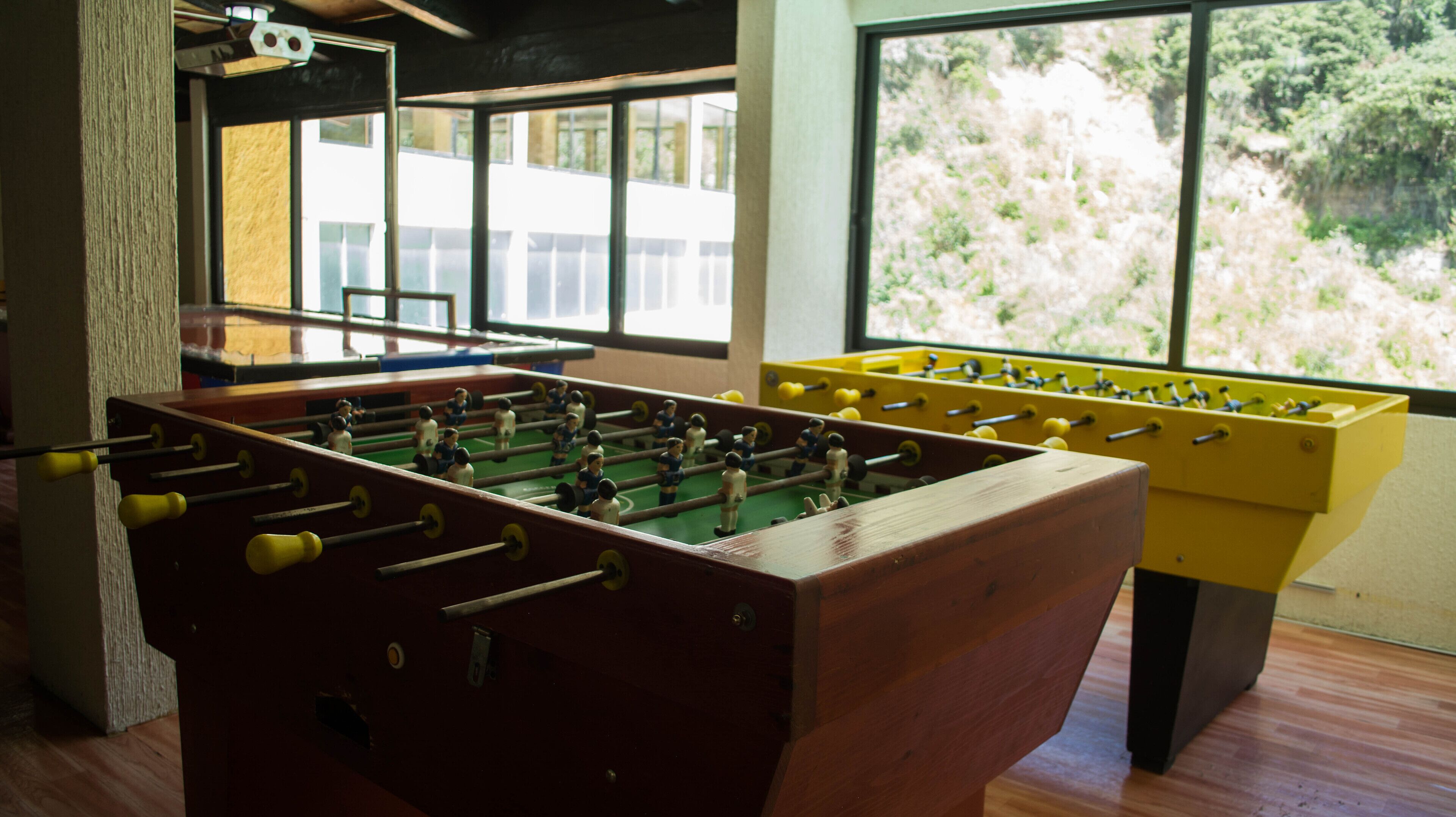 Una habitación con dos mesas de futbolín, una de madera y otra amarilla, y un ventanal con vista a árboles.