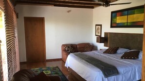 Un dormitorio con cama, sofá, ventilador de techo, una puerta y un cuadro colorido en la pared.