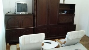 A living room with a dark wooden cabinet, a television, two white armchairs, and a small round table. - Citadela - Large Double Room (Sombor)