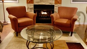 Una sala de estar con chimenea de piedra, dos sillones, una mesa de centro de cristal y una lámpara de pie.