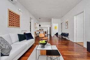 Un salon moderne avec un canapé blanc, une table basse sur laquelle se trouve un saladier de pommes, et une cuisine en arrière-plan.