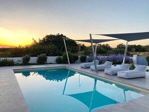 A rectangular swimming pool with a clear blue water surface, surrounded by a light-colored deck. There are two white lounge chairs with cushions under a large, dark-colored canopy. The pool is set against a backdrop of greenery and a clear sky.