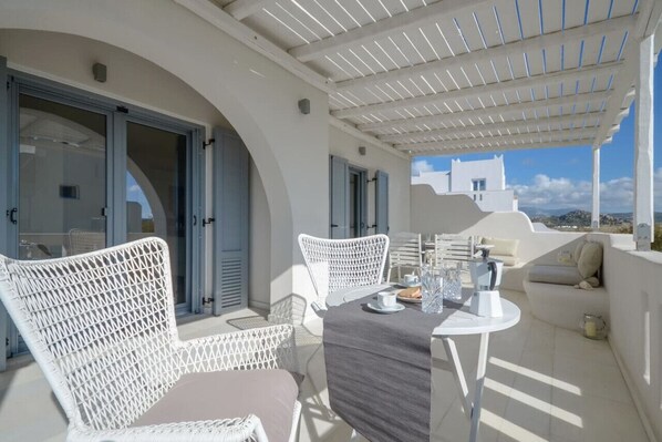 A white wicker chair and table set on a balcony with a pergola overhead, overlooking a coastal town.