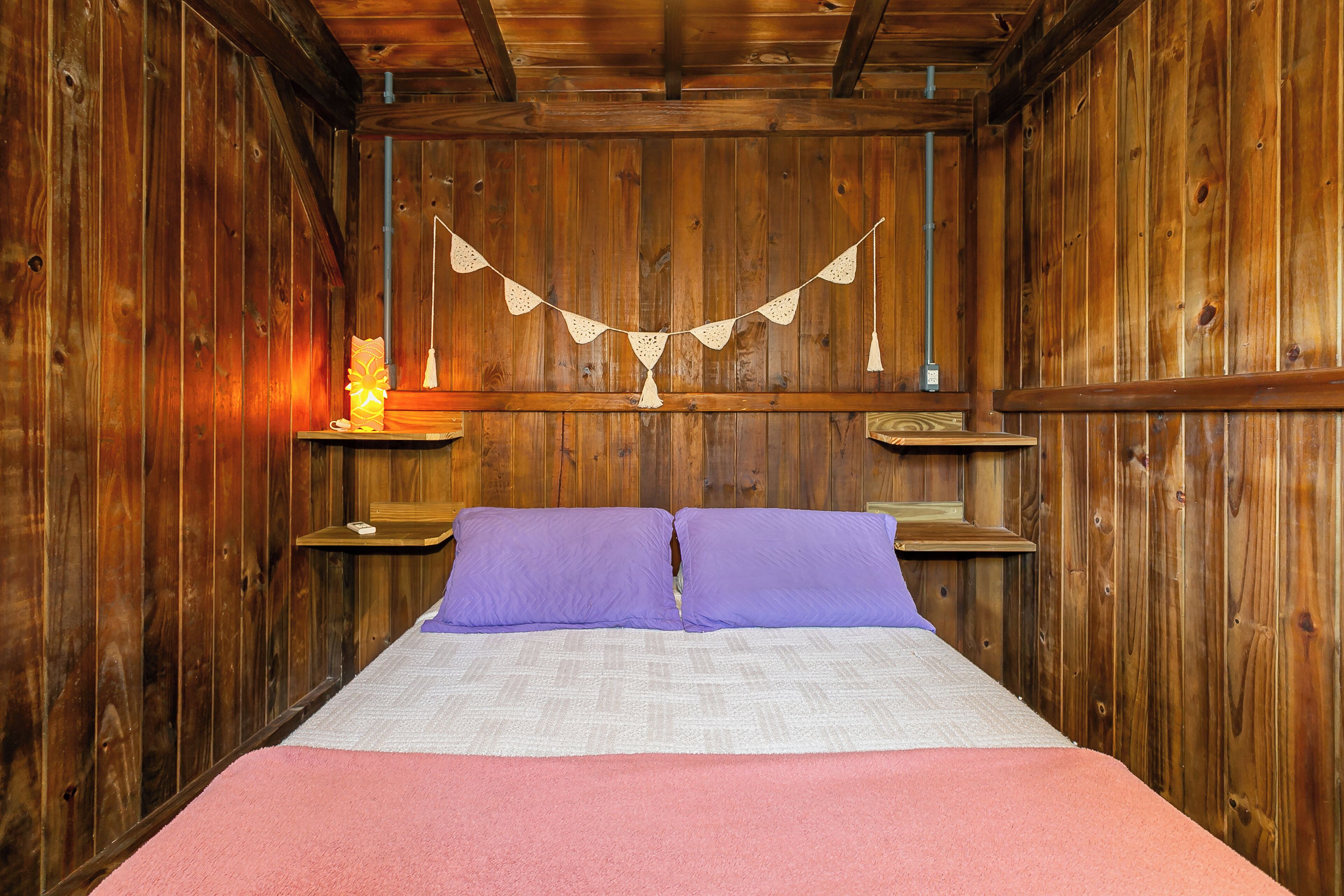A wooden room with a bed, purple pillows, a pink blanket, and a string of lights.