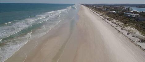A long, sandy beach with waves lapping at the shore, a clear sky, and a distant view of a town with buildings and a body of water.
