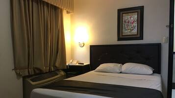 A hotel room with a bed, a bedside table with a phone, a wall-mounted light, and a window with curtains.
