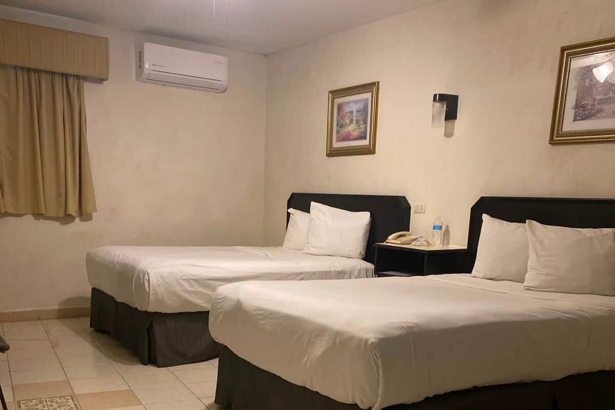 Habitación de hotel con dos camas, un equipo de aire acondicionado, un ventilador de techo, un cuadro enmarco y una mesita de noche con un teléfono y una botella de agua.