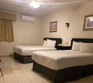 Hotelkamer met twee bedden, een airconditioner, een plafondventilator, een ingelijst schilderij en een nachtkastje met een telefoon en waterfles.