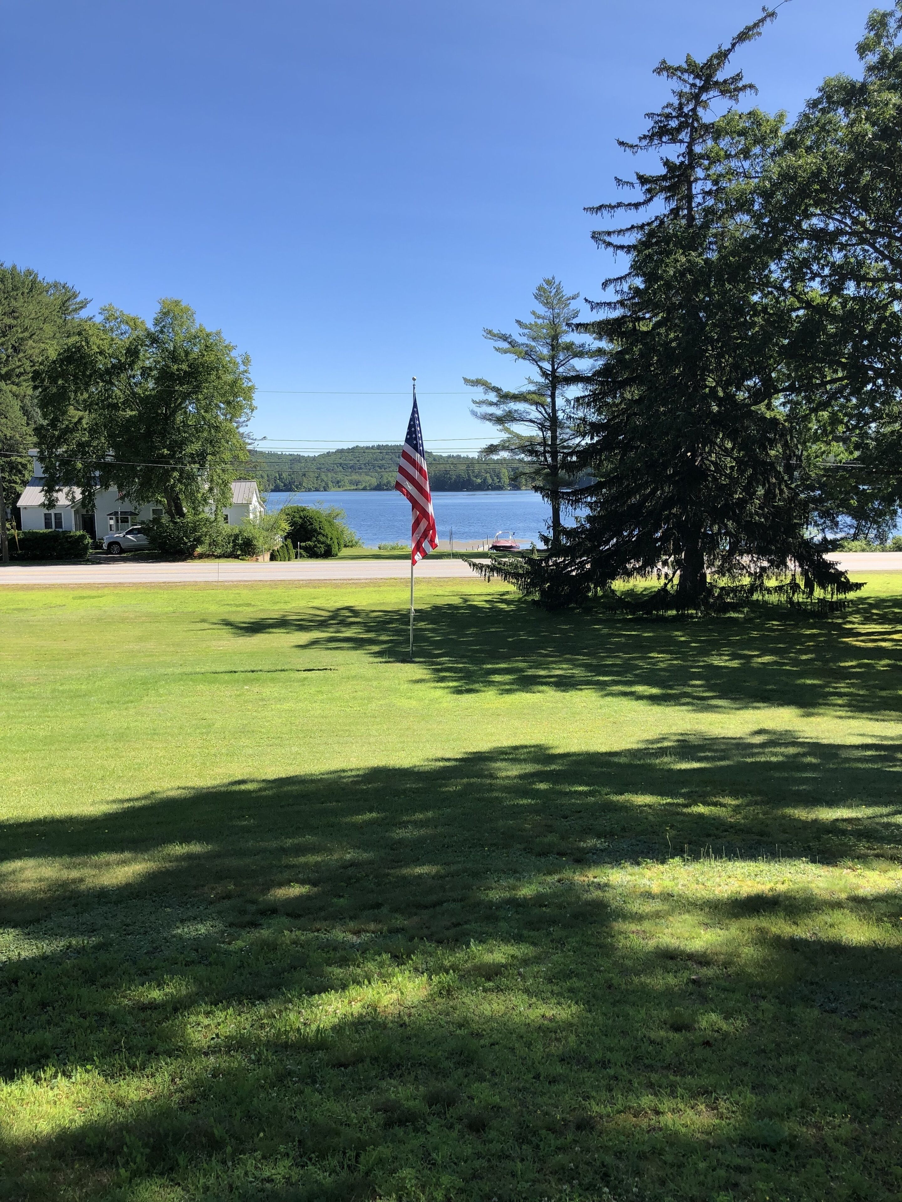 A grassy field with a flagpole and an American flag. There is a large tree to the right and a house in the background.
