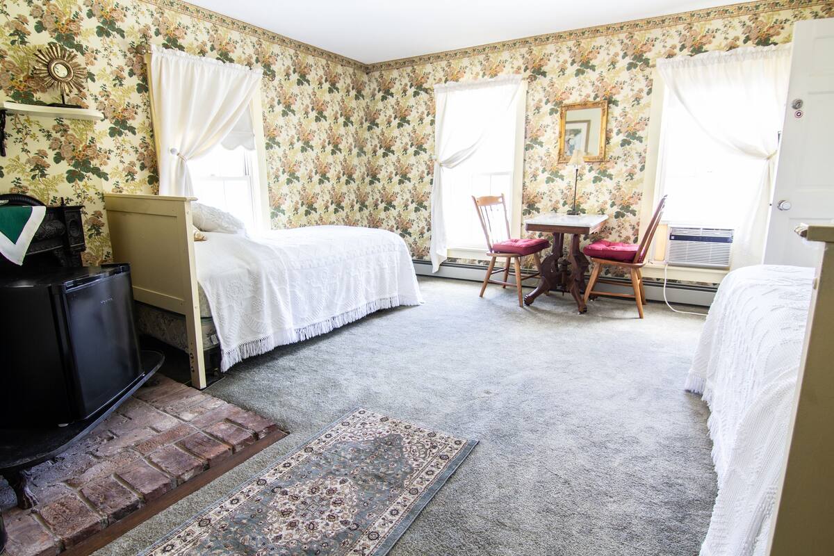 Quarto com duas camas, uma mesinha com cadeira, radiador e tapete com padrão de tijolos.