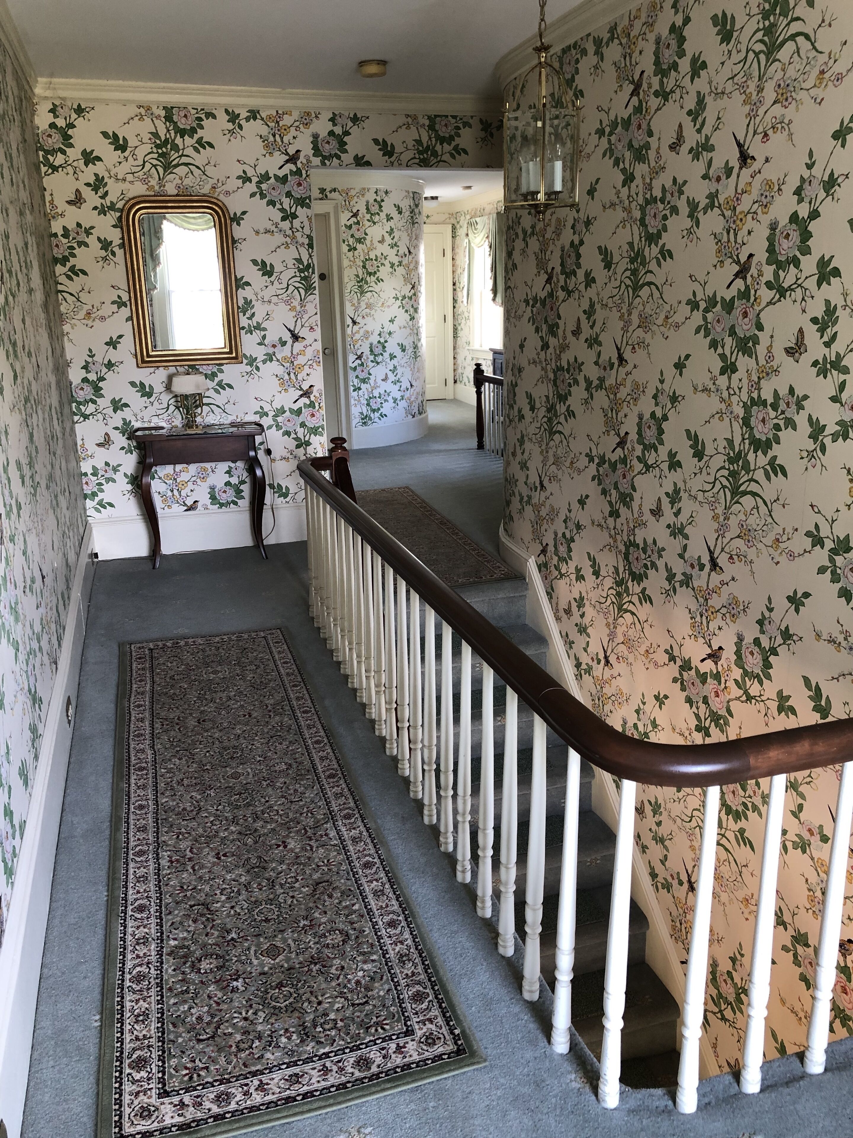 Un pasillo con papel tapiz floral, un banco de madera, un espejo y una escalera con barandillas blancas.