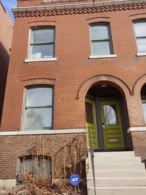 A two-story brick building with a green door, large windows, and a small garden area with dried plants.