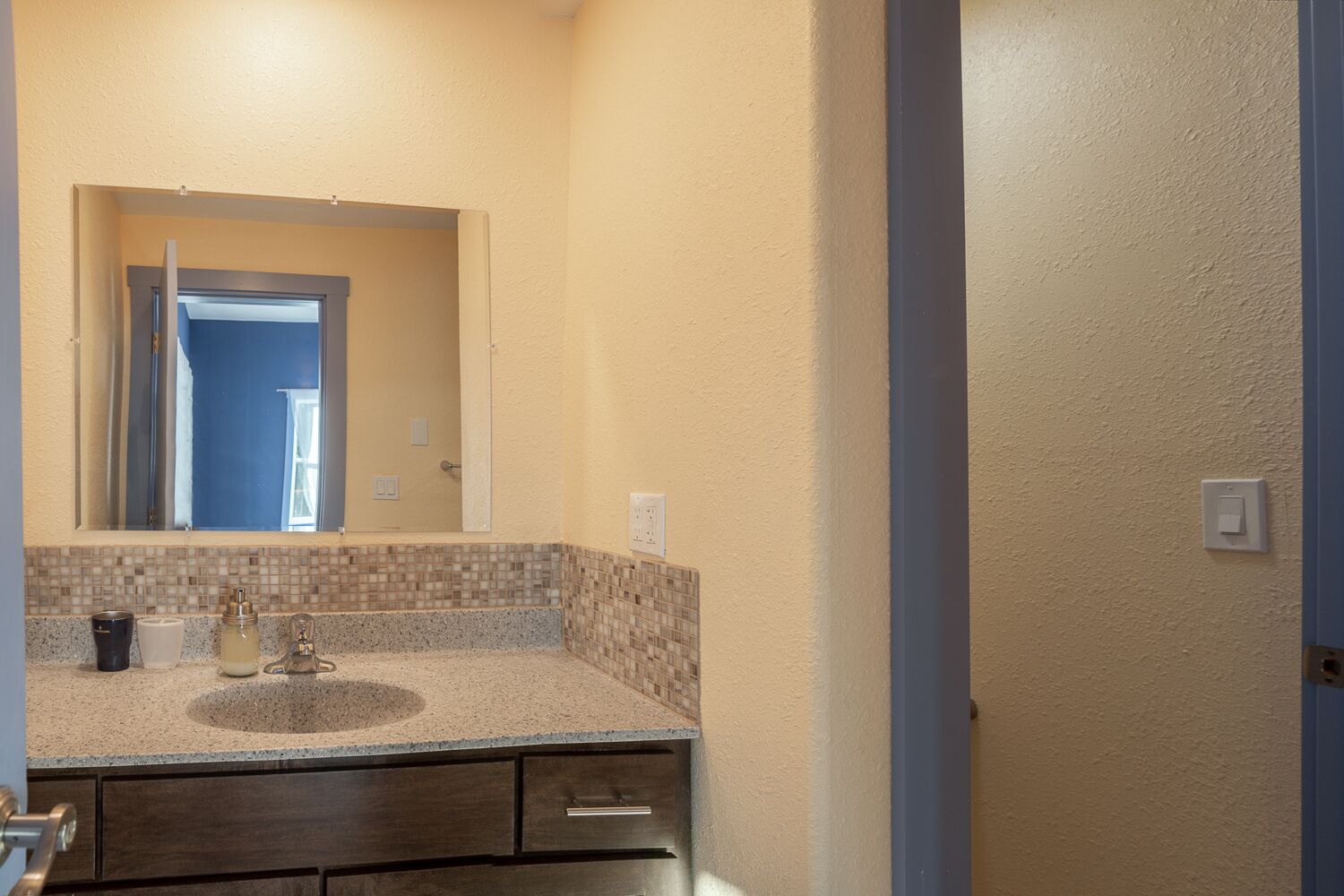 Une salle de bain avec un mur recouvert de carreaux beiges, un lavabo avec un pare-éclaboussures en mosaïque et un miroir au-dessus.