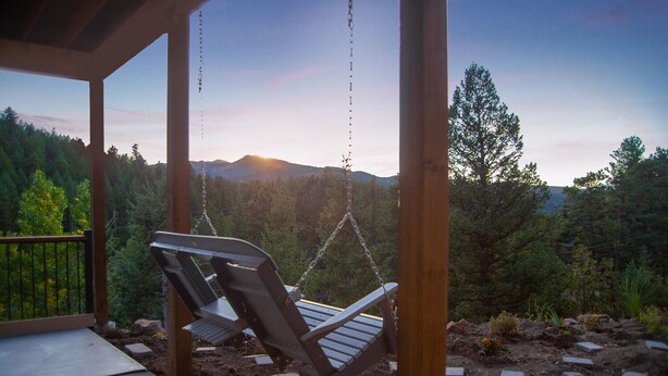 Une terrasse en bois avec un fauteuil suspendu, donnant sur une zone boisée et offrant une vue sur des montagnes en arrière-plan.